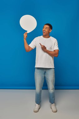 excited african american man in denim jeans pointing at speech bubble on blue backdrop, full length clipart