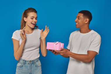Neşeli Afrikalı Amerikalı adam, heyecanlı kız arkadaşına mavi arka planda hediye sunuyor.
