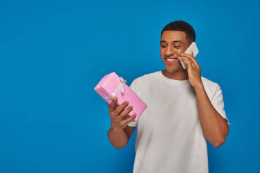 Neşeli Afrikalı Amerikalı adam akıllı telefondan konuşuyor ve mavi arka planda hediyesini tutuyor.