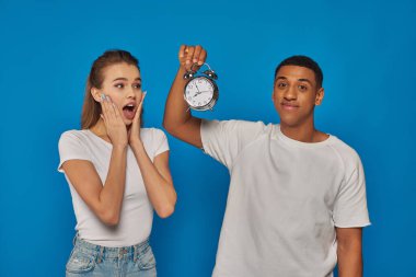 morning time, happy african american man holding alarm clock near astonished woman on blue backdrop clipart