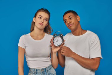 morning time, two displeased interracial couple holding retro alarm clock on blue backdrop clipart