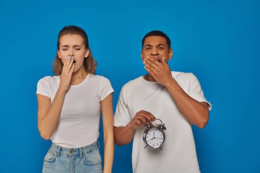 african american man holding alarm clock and yawning with woman on blue backdrop, sleepy couple clipart