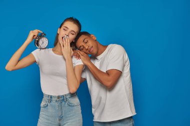 woman holding alarm clock and sleeping with african american man on blue backdrop, tired couple
