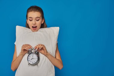 amazed young woman holding pillow and vintage alarm clock on blue background, early morning clipart