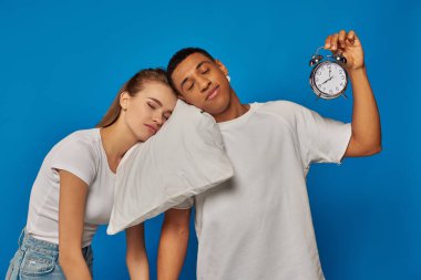 interracial couple sleeping on pillow near vintage alarm clock on blue backdrop, early morning clipart
