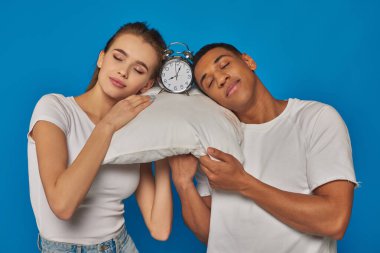 positive interracial couple sleeping together on pillow near alarm clock on blue background clipart