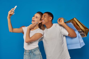 Tüketim, mutlu ırklar arası çiftler alışveriş torbaları tutuyor ve mavi arka planda selfie çekiyorlar.