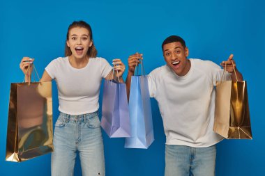 heyecan verici ırklar arası çift ellerinde alışveriş torbalarıyla mavi arka planda kameraya bakıyor, tüketicilik