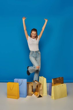 Alışveriş yapmaktan zevk alan kadın, mavi arka planda alışveriş torbalarının yanında el kaldırarak duruyor.