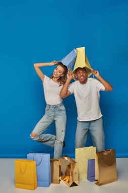 Pozitif çok kültürlü çiftler günlük giysiler içinde ellerinde alışveriş çantalarıyla mavi arka planda, perakende neşesi