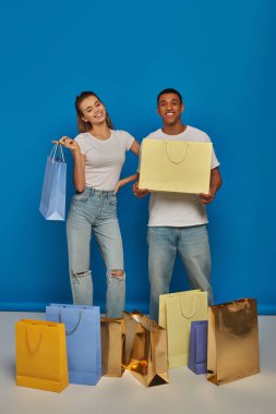 joyful multiethnic couple in casual attire holding shopping bags on blue backdrop, buying spree clipart