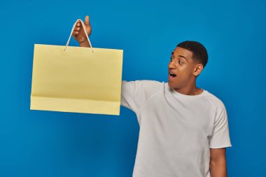 excited african american man in casual attire holding shopping bag on blue backdrop, retail joy clipart
