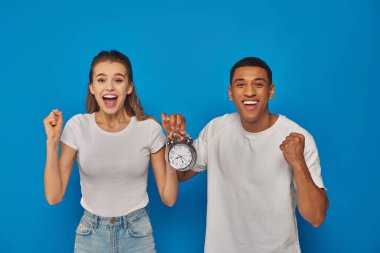 excited interracial couple with open mouth holding alarm clock on blue background, face expression clipart