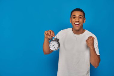 excited african american man with open mouth holding alarm clock on blue background, face expression clipart