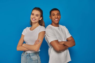 cheerful multicultural couple standing with folded arms and looking at camera on blue background clipart