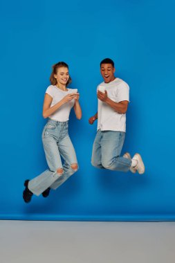 happy multicultural couple using smartphones and jumping  on blue background, full length view clipart