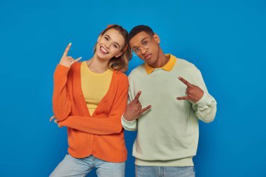 Şık ve genç ırklar arası çift, günlük giysiler içinde, mavi arka planda el kol hareketi yapıyorlar, kaya işareti gösteriyorlar.