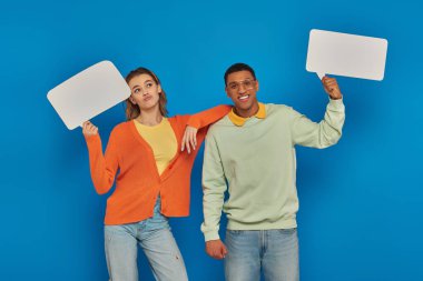 cheerful interracial couple in smart casual clothes holding blank speech bubbles on blue backdrop clipart