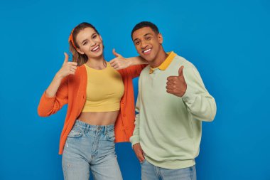 happy multicultural couple looking at camera and showing thumbs up on blue backdrop, like gesture clipart