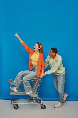 positive african american man riding shopping cart with girlfriend inside of it on blue background clipart