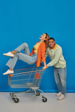 cheerful african american man riding shopping cart with girlfriend inside of it on blue background clipart