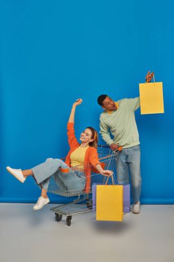 Alışveriş arabasındaki mutlu Afro-Amerikan erkek ve genç kadın mavi arka planda alışveriş torbaları taşıyorlar.