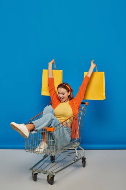 Heyecanlı genç bir kadın alışveriş arabasında oturuyor ve mavi arka planda alışveriş torbaları tutuyor.