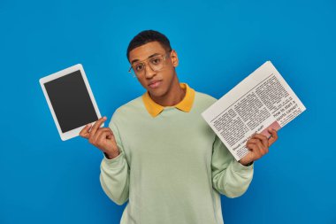 handsome african american man in eyeglasses holding digital tablet and newspaper on blue backdrop clipart