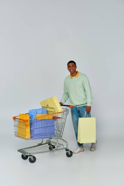 Heyecanlı Afro-Amerikalı adam tramvay ve alışveriş çantalarıyla gri arka planda duruyor, tüketicilik