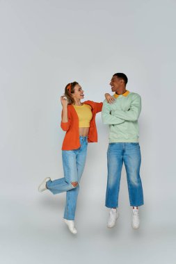 happy young woman sticking out tongue and looking at african american man on grey backdrop, levitate clipart