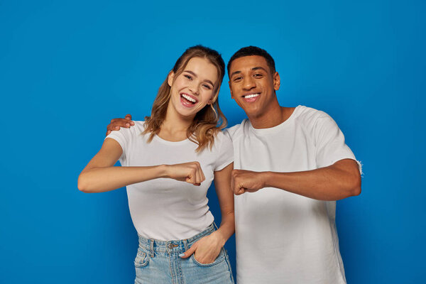 happy interracial couple fist bumping on blue backdrop, african american man and excited woman