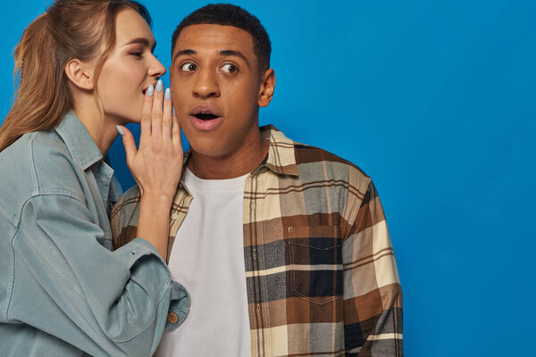 young woman whispering in ear of amazed african american on blue background, sharing secrets
