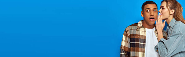young woman whispering in ear of amazed african american on blue background, sharing secrets, banner