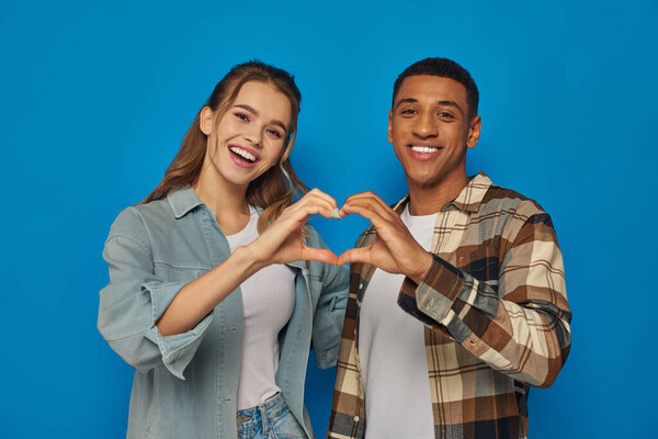 cheerful interracial couple showing heart sign with hands and looking at camera on blue background