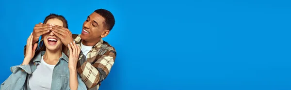 stock image excited african american man covering eyes of woman with open mouth on blue background, banner