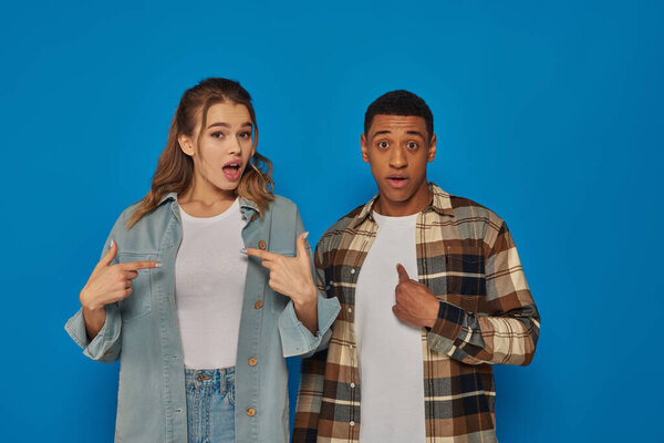 astonished interracial couple with open mouth pointing at themselves with fingers on blue background