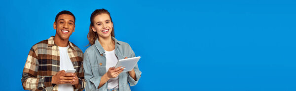 счастливая межрасовая пара, держащая смартфон и цифровой планшет на синем фоне, баннер