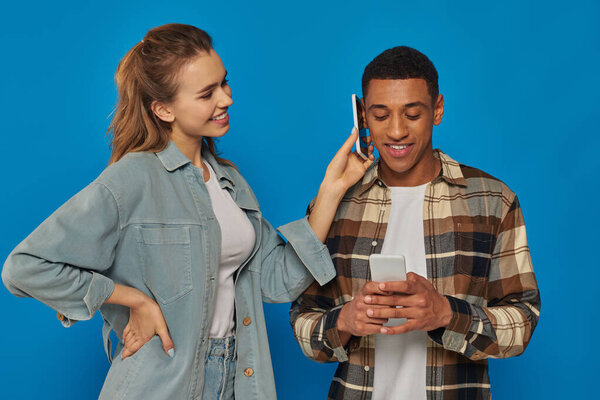 happy woman holding smartphone near ear of african american guy on blue background, communication