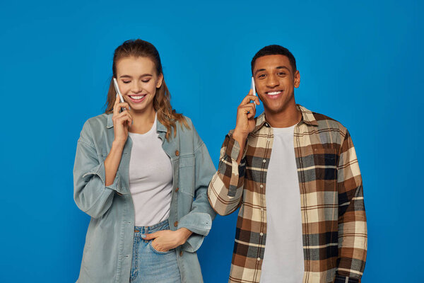 cheerful woman and happy african american man having phone call, using smartphones on blue backdrop