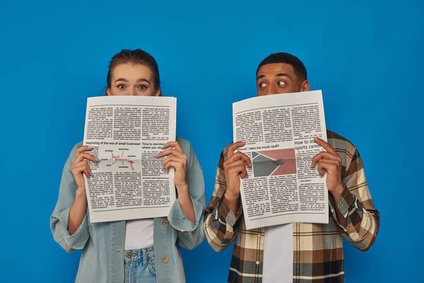 межрасовые мужчина и женщина скрывают лица с газетами на синем фоне, читая новости