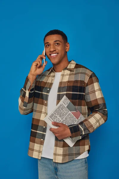 stock image happy african american man in plaid shirt having phone call and holding newspaper on blue backdrop