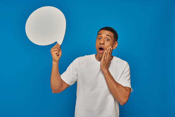 stunned african american man with open mouth holding blank speech bubble on blue background