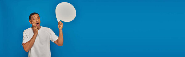 stunned african american man with open mouth holding blank speech bubble on blue background, banner