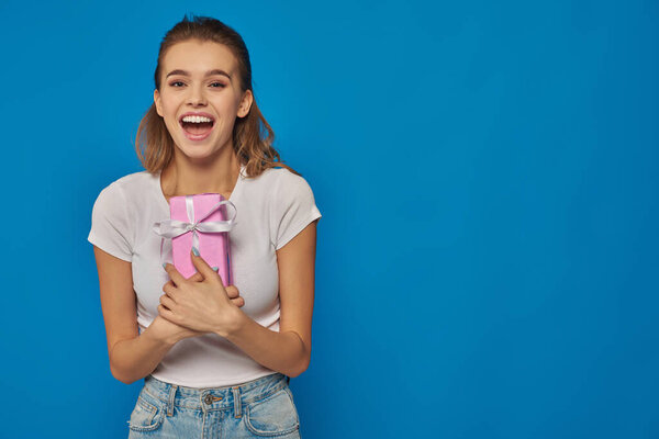 excited young woman holding gift box and looking at camera on blue background, festive occasions
