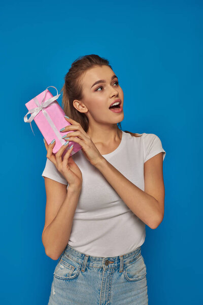 amazed young woman holding gift box and looking away on blue background, festive occasions