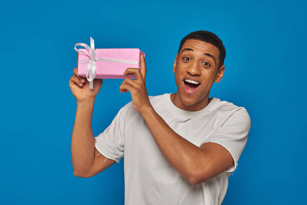 возбужденный африканский американец держит завернутый подарок на голубом фоне, праздничные мероприятия