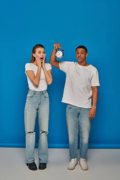 morning time, african american man holding alarm clock near astonished woman on blue backdrop