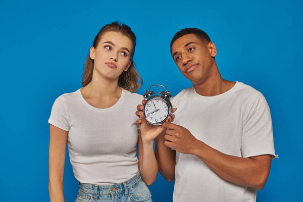 morning time, two displeased interracial couple holding retro alarm clock on blue backdrop