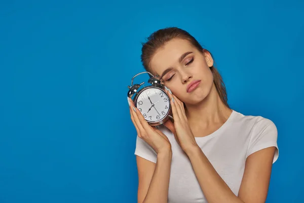 sleepy young woman with closed eyes holding retro alarm clock in hands on blue backdrop, morning time