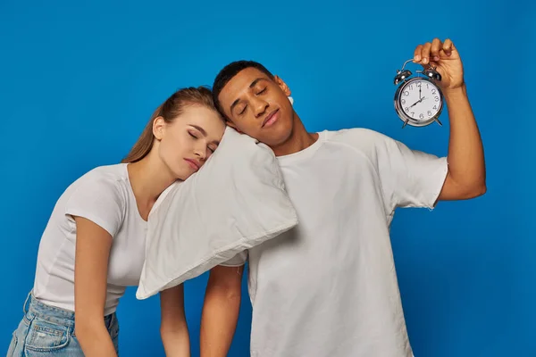 interracial couple sleeping on pillow near vintage alarm clock on blue backdrop, early morning